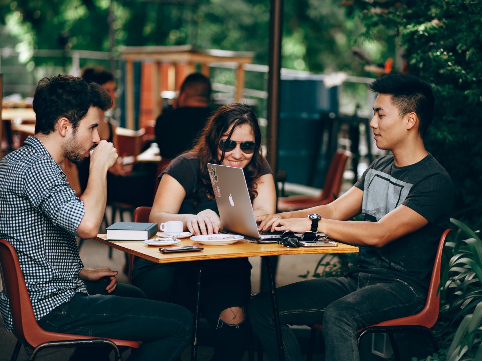 Two young men and a young woman sit outside of a cafe working on laptops.