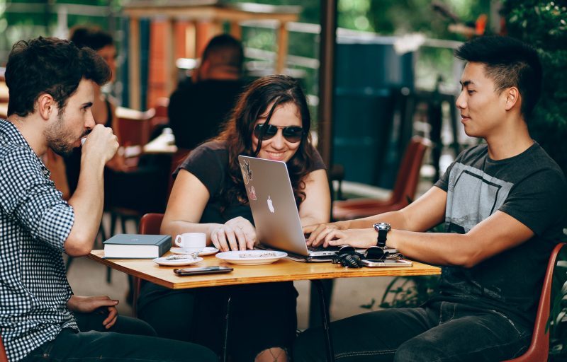 Two young men and a young woman sit outside of a cafe working on laptops.