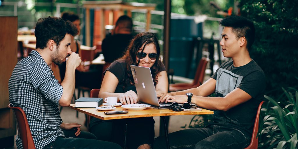 Two young men and a young woman sit outside of a cafe working on laptops.