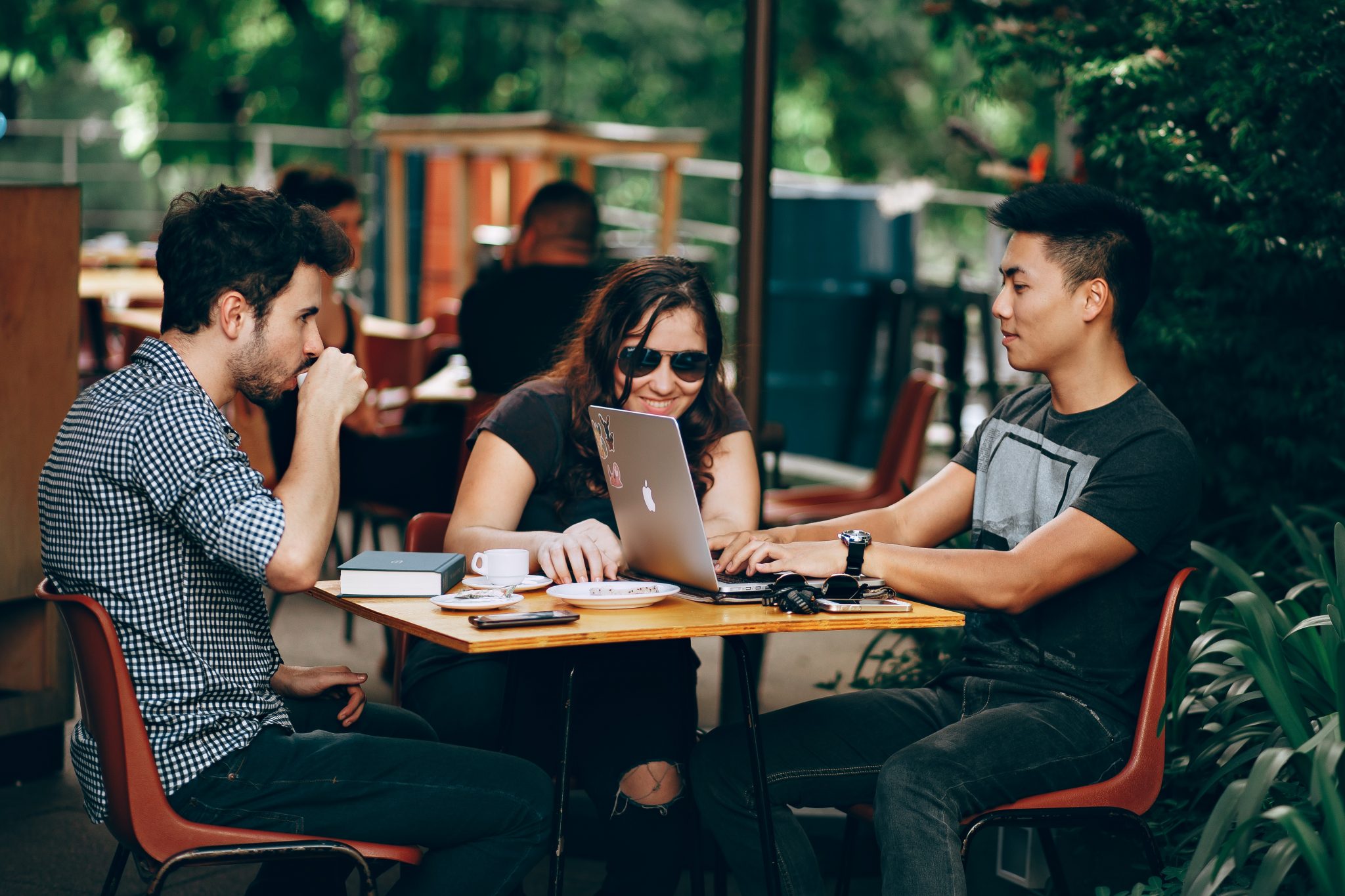 Two young men and a young woman sit outside of a cafe working on laptops.