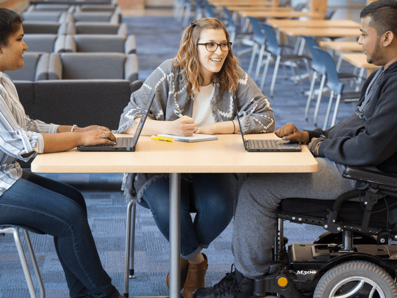 Three students working in front of laptops in a library setting.