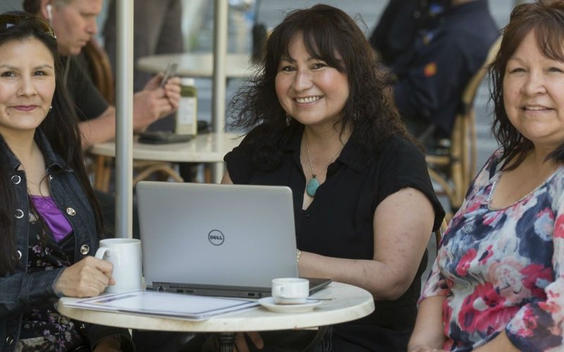 Three indigenious women smiling and sitting in front of a laptop in a cafe setting.