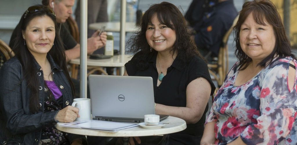 Three indigenious women smiling and sitting in front of a laptop in a cafe setting.