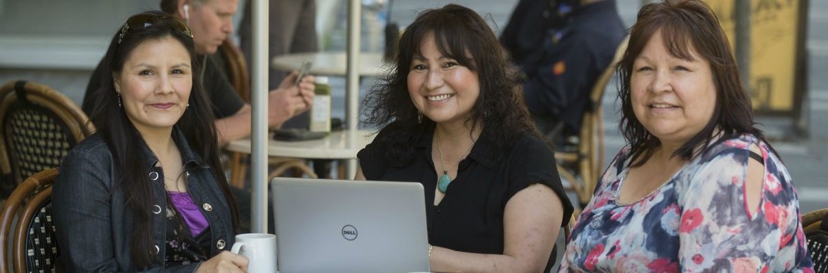 Three indigenious women smiling and sitting in front of a laptop in a cafe setting.