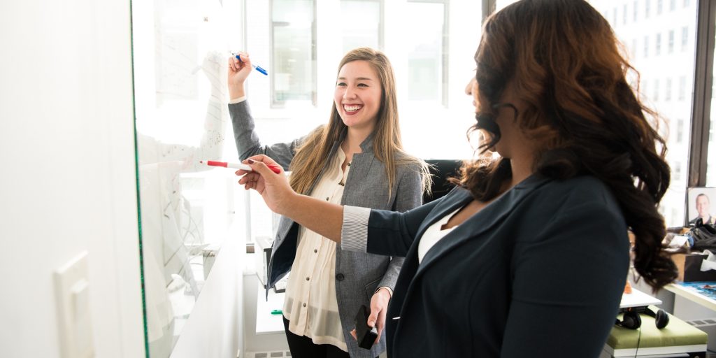 Two women smiling while writing on a whiteboard.