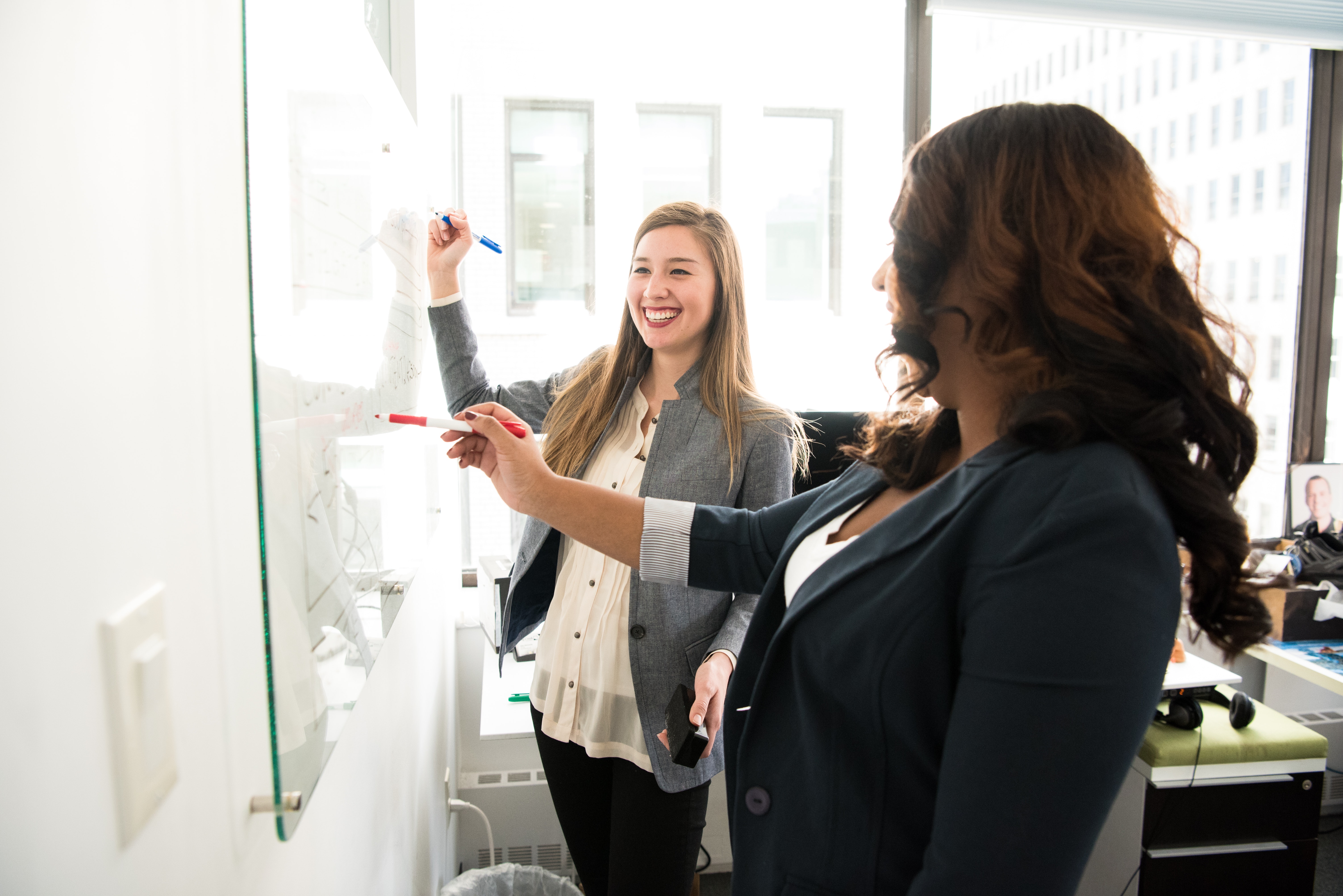 Two women smiling while writing on a whiteboard.