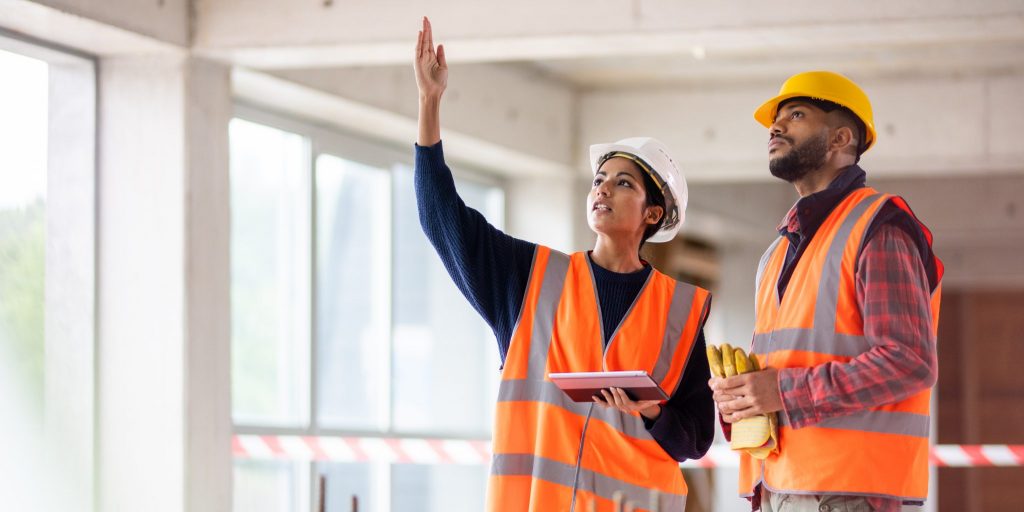 Photo of a woman of Indian ethnicity holding a digital tablet and talking to a young construction worker while pointing at something at the construction site.