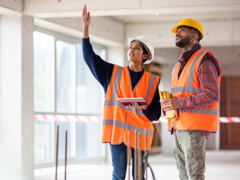 Photo of a woman of Indian ethnicity holding a digital tablet and talking to a young construction worker while pointing at something at the construction site.