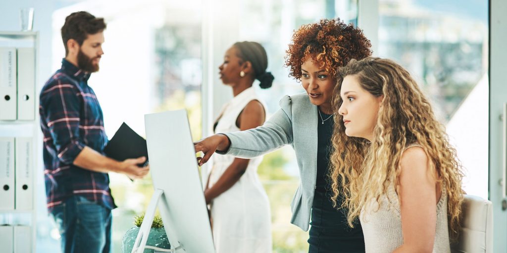 Shot of two businesswomen working together on a computer in an office with their colleagues in the background