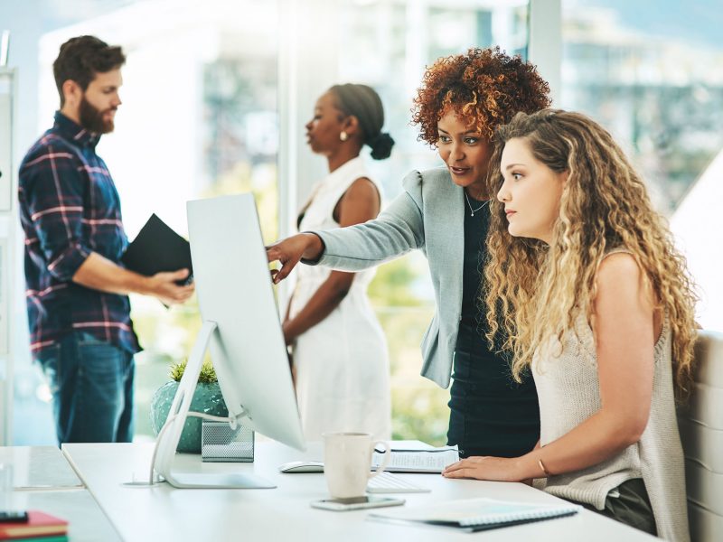 Shot of two businesswomen working together on a computer in an office with their colleagues in the background