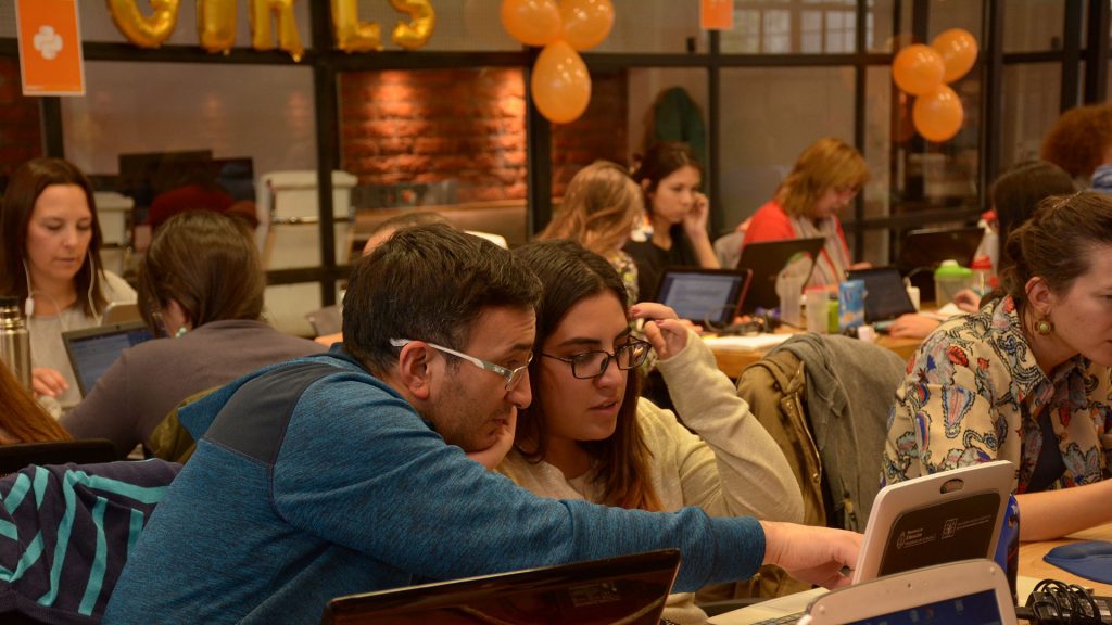 Two people working together at a laptop in an open workspace with others