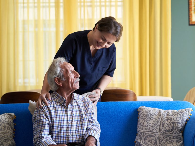 Standing care worker leaning in to smile at senior seated on a couch