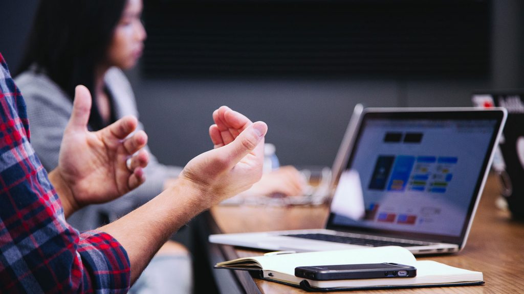 Hands gesticulating in front of open laptop; person on laptop in background