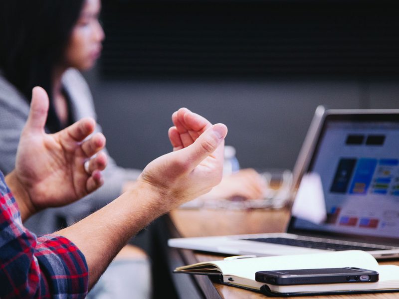 Hands gesticulating in front of open laptop; person on laptop in background