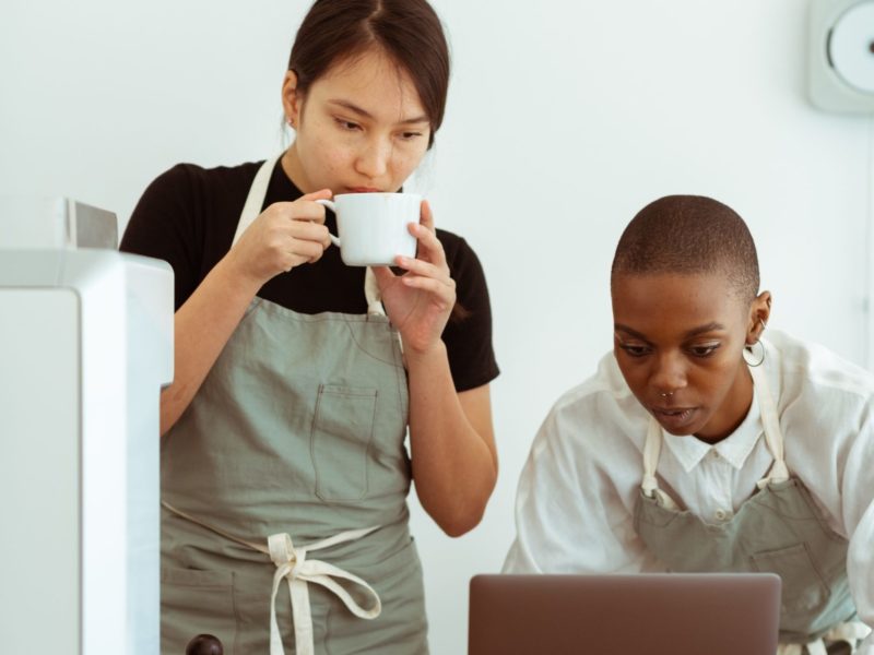 Two woman, one sipping coffee, both are looking at a laptop screen
