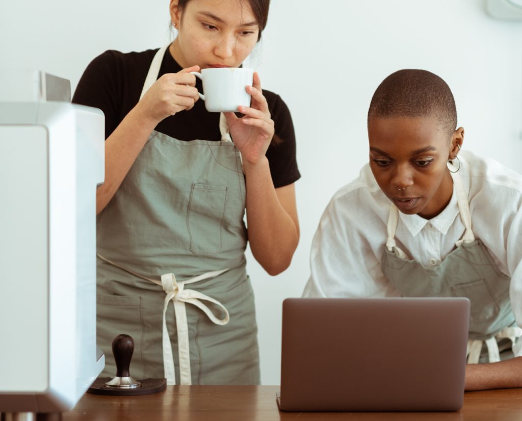 Two woman, one sipping coffee, both are looking at a laptop screen