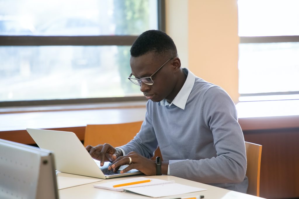 Young student using a laptop and taking notes in a library.