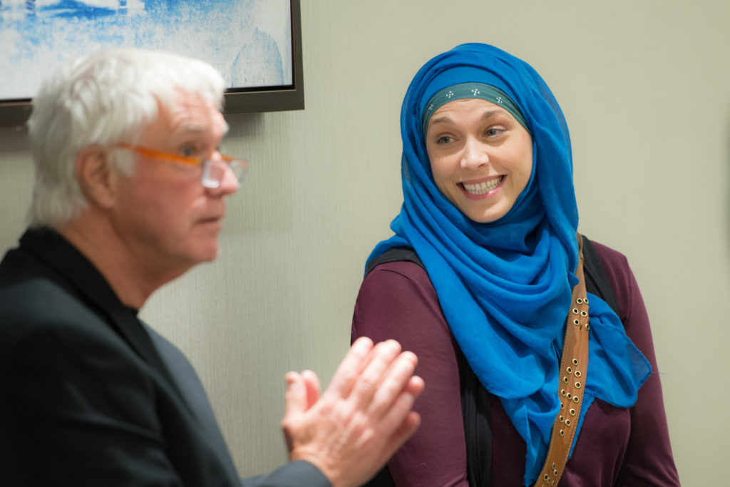 Man speaking to a group while woman listens smiling.