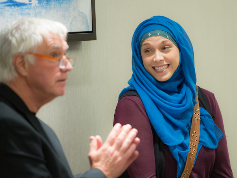 Man speaking to a group while woman listens smiling.