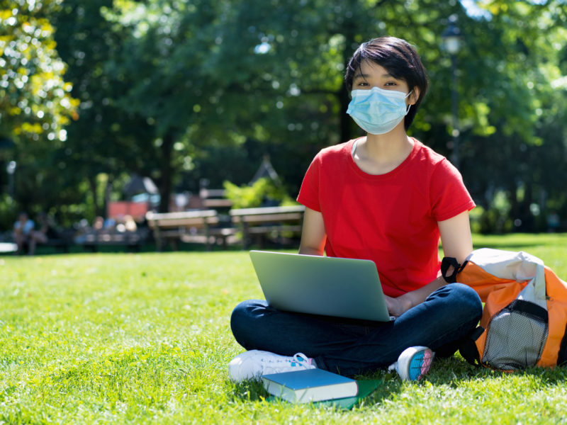 Student sitting outside of university campus with a clinical mask on. 
