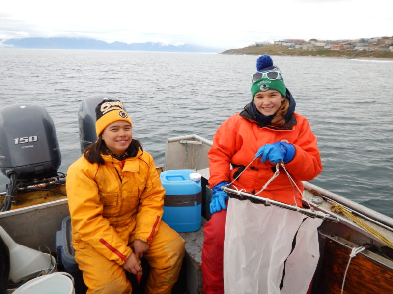 Two young individuals in a boat holding up a plankton net.