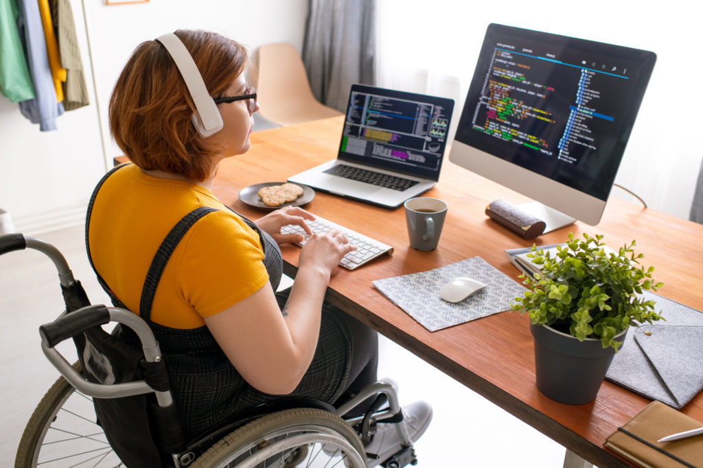 Woman in wheelchair working from home looking at dual computer screens.