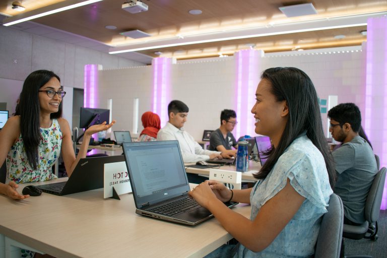 Students in a classroom sit across from eachother with laptops.