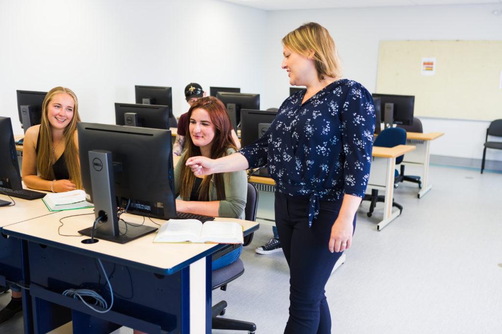 Students in a computer lab with teacher pointing at screen.