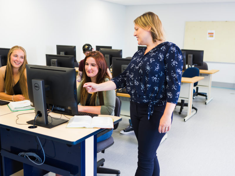 Students in a computer lab with teacher pointing at screen.