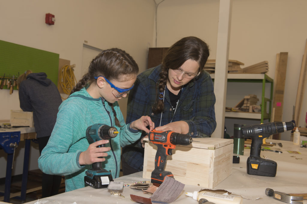 A woman teaching a younger girl how to drill and build a wooden box.
