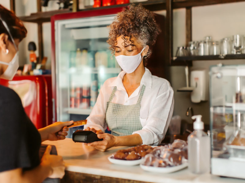 Business owner in a face mask taking a card payment from a customer. Mature businesswoman serving a customer over the counter in her cafe. Customer service during the COVID-19 pandemic.