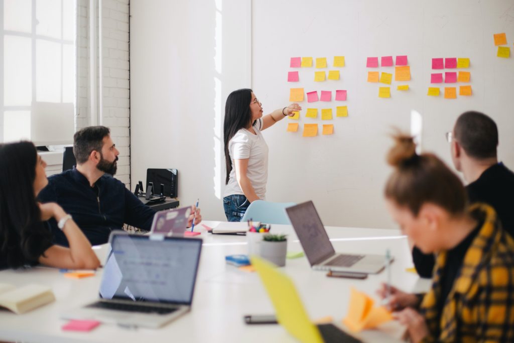 A group of people sitting around a table with a person in a white t-shirt presenting and pointing to a series of pink, yellow and orange sticky notes on a wall. 