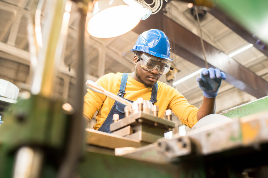 Serious busy young black factory engineer in hardhat and safety goggles examining milling lathe and repairing it while working at production plant