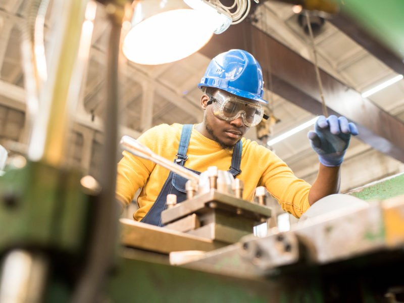 Serious busy young black factory engineer in hardhat and safety goggles examining milling lathe and repairing it while working at production plant