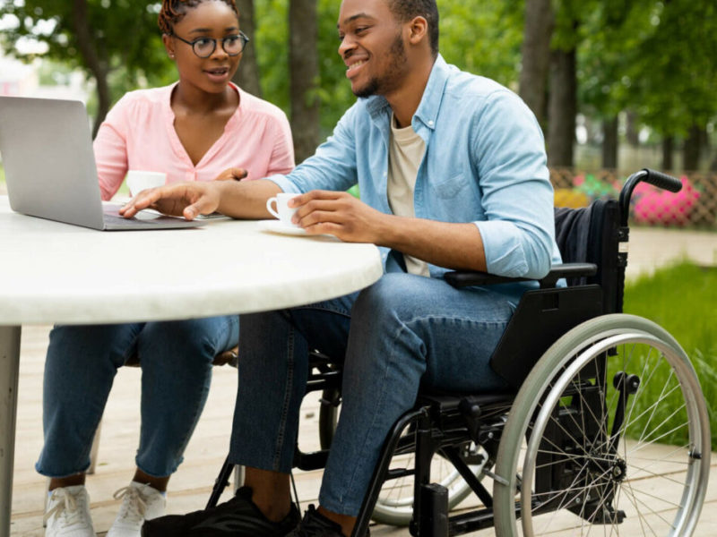 man in a wheelchair looking at a computer and talking to a friend