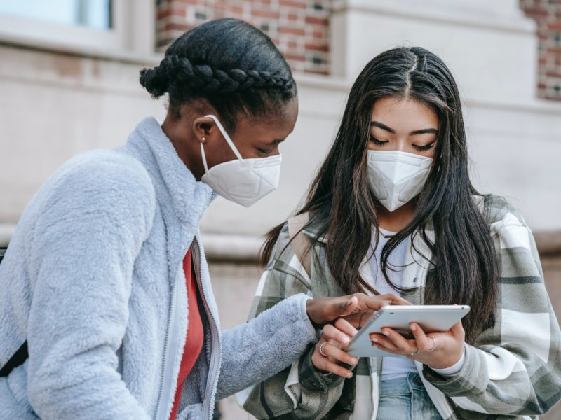 Two people in masks are standing outside a building, looking at a tablet.