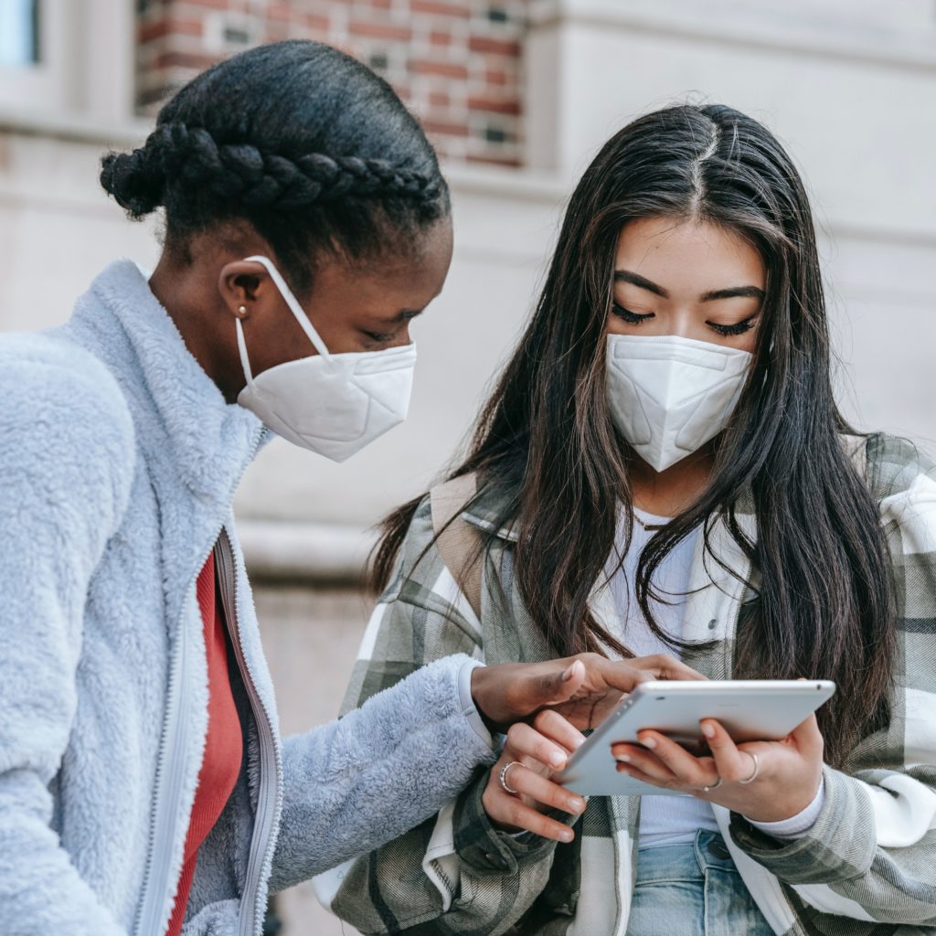 Two people in masks are standing outside a building, looking at a tablet. 