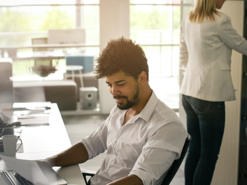 A person is working at a desk, reviewing paperwork while someone is opening a cabinet door behind them