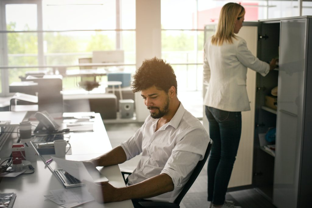 A person is working at a desk, reviewing paperwork while someone is opening a cabinet door behind them