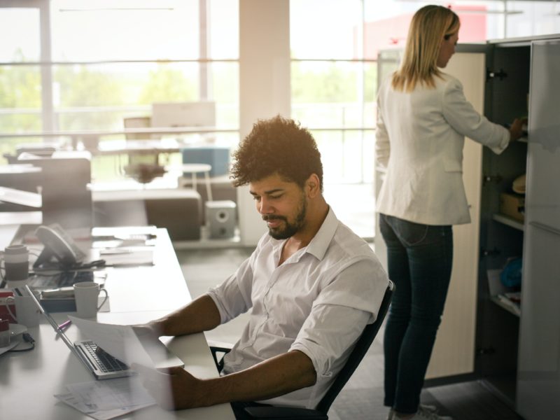 A person is working at a desk, reviewing paperwork while someone is opening a cabinet door behind them