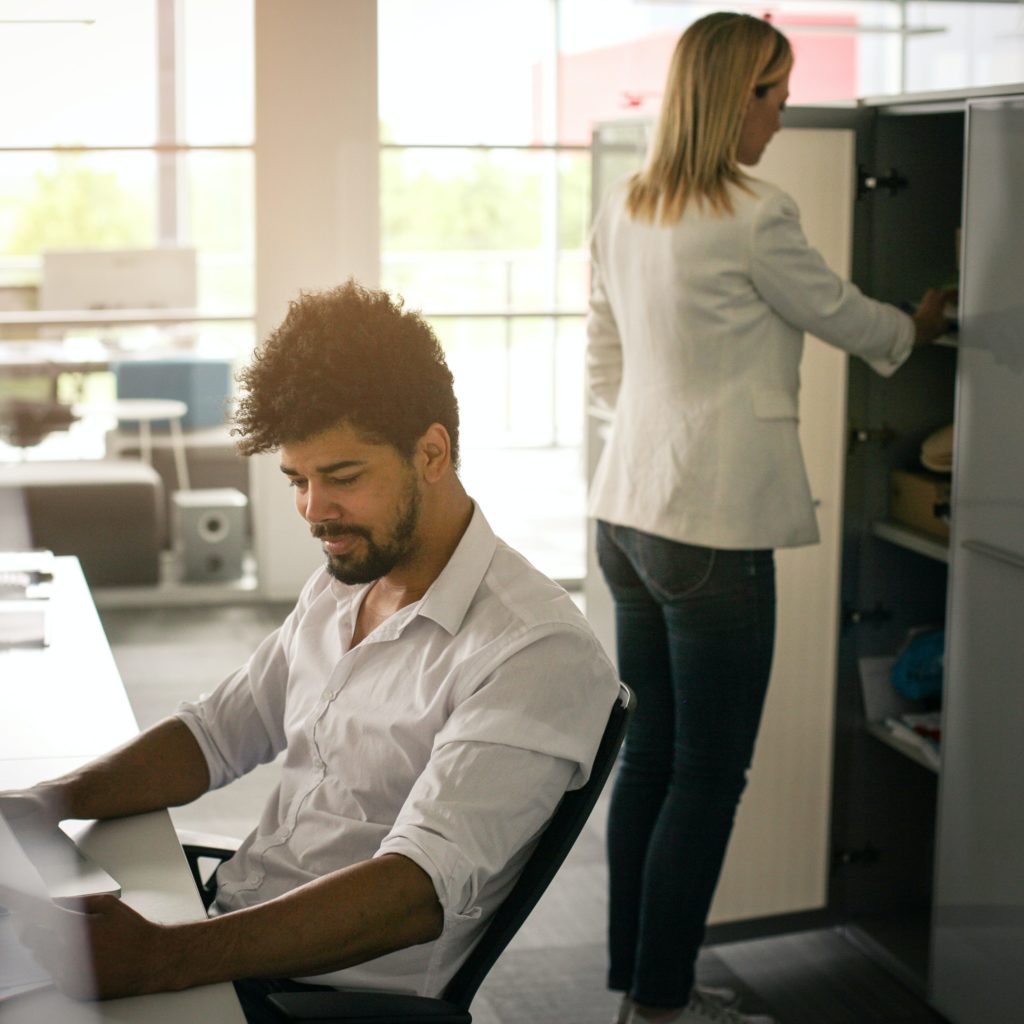 A person is working at a desk, reviewing paperwork while someone is opening a cabinet door behind them