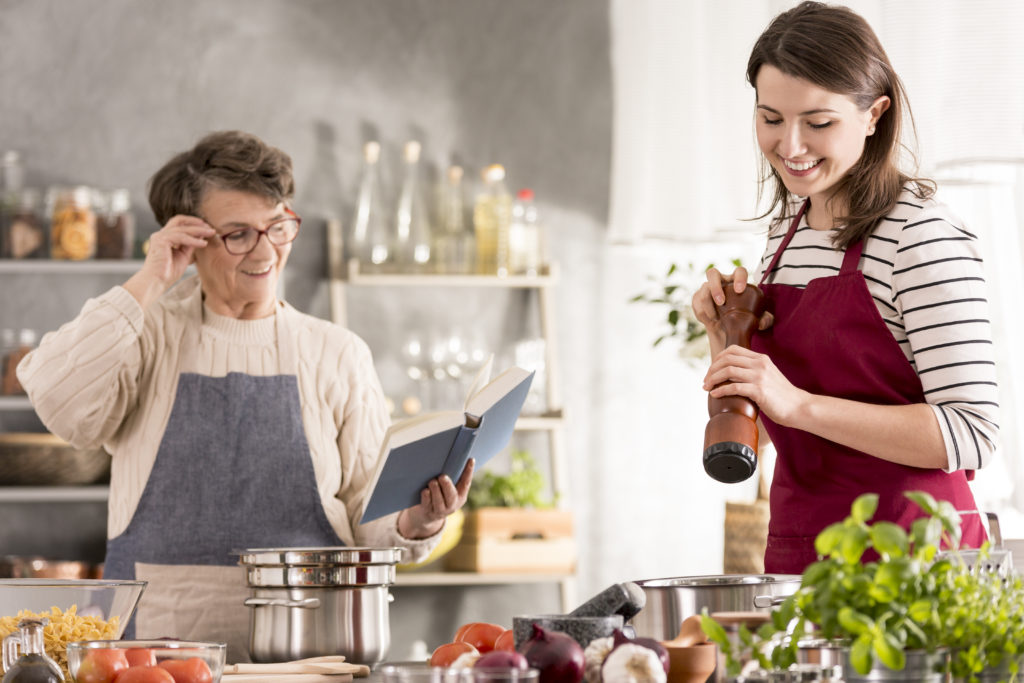 Woman cooking and her grandmother reading recipe from cookbook