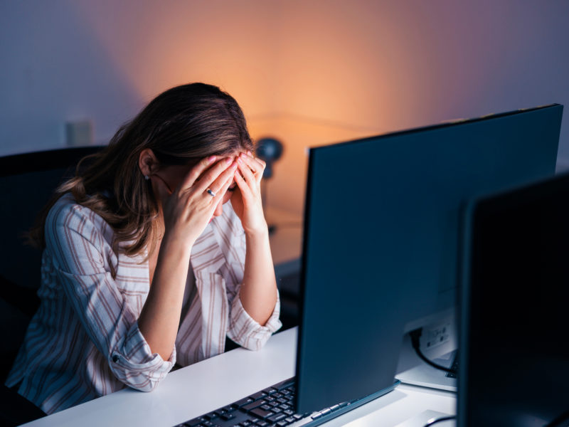 Tired and stressed out woman working late in an office, leaning at her desk and holding head in hands; anxious woman working overtime