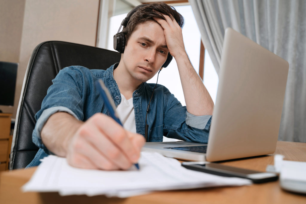 Tired young man working at home office on new project, frustrated guy sitting at his desk writing on papers, thinking on difficult task.