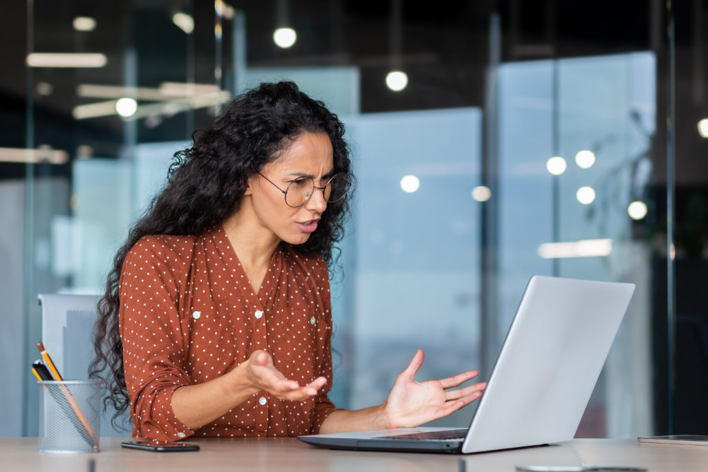 Frustrated business woman looking at laptop screen, dissatisfied female worker spreading hands inside office, latin american woman working at desk using computer for video call online communication.