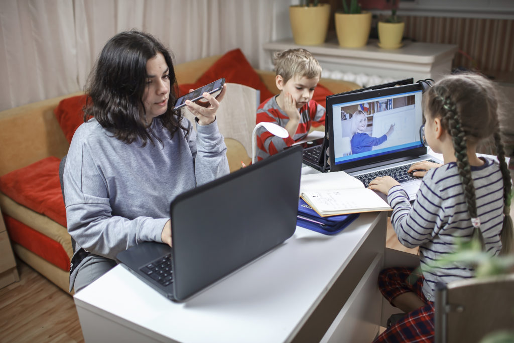 Distant education and work at home, children doing homework, mother working and help them. Elementary school kids during online class with parent working remotely in one room, lockdown, focus on woman