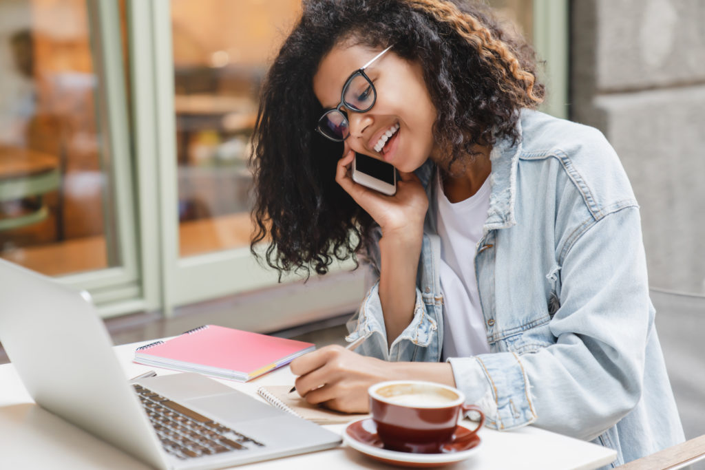 Young successful african-american businesswoman student working on laptop remotely talking on smart phone with colleagues boss, dealing with business partenrs in cafe. Freelance job occupation.