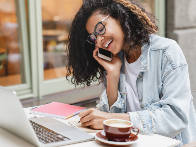 Young successful african-american businesswoman student working on laptop remotely talking on smart phone with colleagues boss, dealing with business partenrs in cafe. Freelance job occupation.