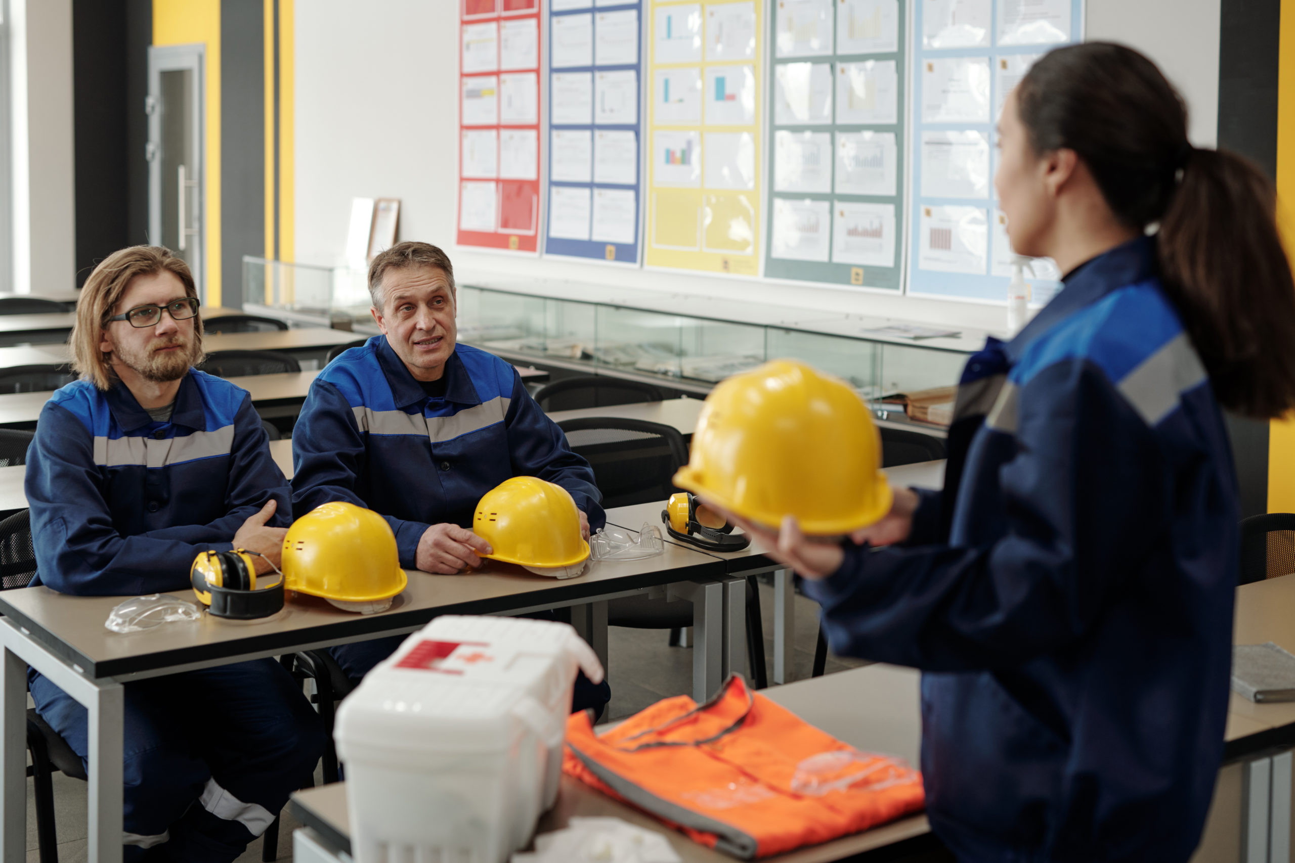 Female instructor holding hardhat and explaining occupational safety rules to workers sitting at table