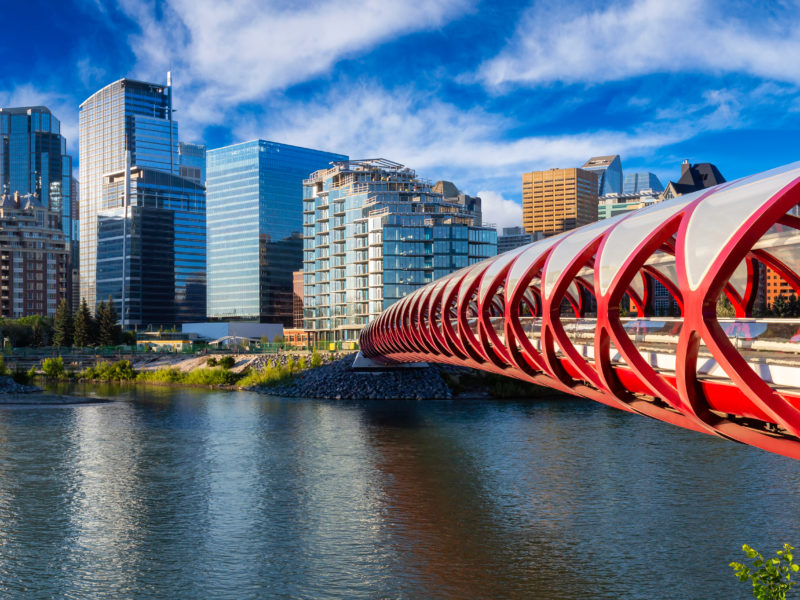 Peace Bridge across Bow River with Modern City Buildings in Background during a vibrant summer sunrise. Taken in Calgary, Alberta, Canada.